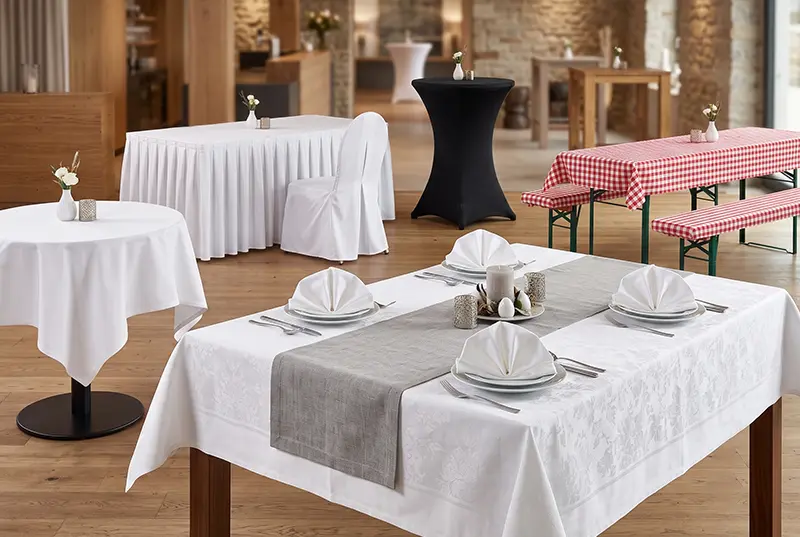 Vielfältige Tischwäsche für die Gastronomie: Elegante weiße Tischdecken mit Servietten, graue Tischläufer, schwarze Stehtischhussen, Stuhlhussen und karierte Biertischdecken in einem modernen Restaurant-Ambiente.